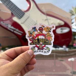 Op de afbeelding: Een kleurrijke sticker met Dr. Teeth and The Electric Mayhem. De sticker toont de bandleden met muziekinstrumenten en de bandnaam. Op de achtergrond is een grote rode en witte gitaar te zien.