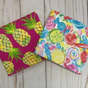 May include: Two square fabric napkins. One is fuchsia with a repeating pattern of yellow and green pineapples. The other is white with a colorful pattern of candies, lemons, and limes.