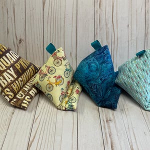 May include: Five different fabric wedge pillows. The pillows are all triangular in shape and have different patterns. One pillow has a brown and white pattern with the words "Old Bay Square" on it. Another pillow has a yellow and white pattern with bicycles on it. Another pillow has a blue and white pattern with a floral design. Another pillow has a light blue and white pattern with stripes. The last pillow has a light blue and white pattern with a floral design.