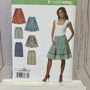 May include: Simplicity sewing pattern featuring six skirt designs. The pattern includes sizes 12-20. The image shows a woman wearing a skirt similar to one of the designs. The pattern is displayed on a white wooden stand.