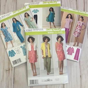 May include: Several Simplicity sewing pattern envelopes are displayed, showcasing various dress and outfit designs. The patterns feature images of the finished garments, with models wearing dresses, tops, and pants in different colors and styles. The envelopes have the Simplicity logo.