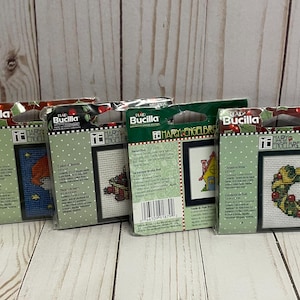 May include: Five different cross-stitch patterns from Mary Engelbreit. The patterns are on white cardstock with colorful illustrations and text. The patterns include a gnome, a wreath, a house, a snowman, and a stocking.