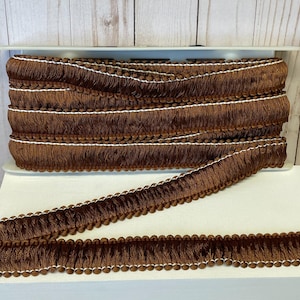 May include: A roll of brown fringe trim with a white accent. The trim features a row of small, round beads along the edge and a fringe of soft, textured material. The trim is displayed on a white surface.