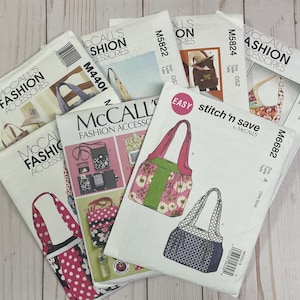 May include: A collection of McCall's Fashion Accessories sewing patterns. The patterns feature illustrations of various bags, including tote bags and handbags. The patterns have a white background with colorful illustrations and text. The patterns are arranged on a light wood surface.