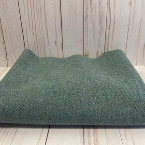 May include: A folded piece of fabric in a muted sage green color. The textile features a dense, textured weave with subtle variations in tone, creating a consistent, speckled appearance across the entire surface of the material.