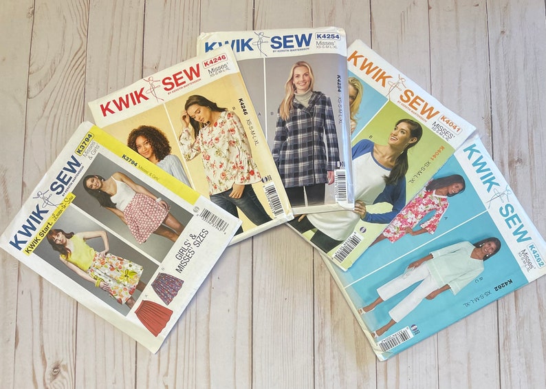 May include: Five different sewing patterns for women's clothing. The patterns are from the brand Kwik Sew and feature different styles, including a blouse, a skirt, a jacket, and pants. The patterns are printed on white paper with colorful illustrations and text.