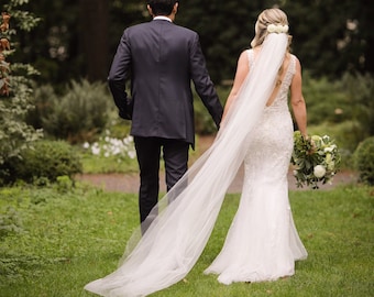 May include: A bride and groom walking away from the camera in a lush green garden setting. The bride is wearing a white wedding dress with a long veil and carrying a bouquet of white flowers. The groom is wearing a black suit.