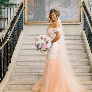 May include: A bride in a pink and white wedding dress with a veil and a bouquet of white and pink flowers stands on a marble staircase.