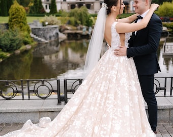 May include: A bride and groom embrace in front of a large building with a pond in the foreground. The bride is wearing a white lace wedding dress with a long train and a veil. The groom is wearing a black suit and a white shirt.