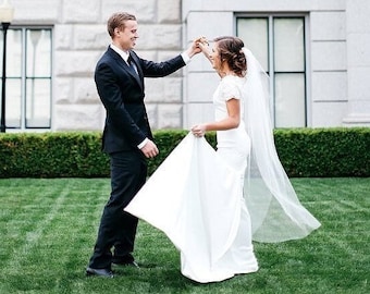 May include: A bride and groom are dancing in front of a white building. The bride is wearing a white wedding dress with a long veil. The groom is wearing a black suit. They are both smiling.