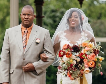 Op de afbeelding: Een bruid in een witte kanten bruidsjurk loopt met haar vader de gang door. Ze draagt ​​een groot boeket met rode, oranje en gele bloemen. Het paar loopt onder een prieel met een rood dak.