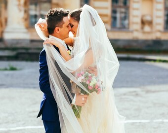 May include: A bride and groom embrace, their faces hidden behind a white veil. The bride holds a bouquet of pink and white flowers.