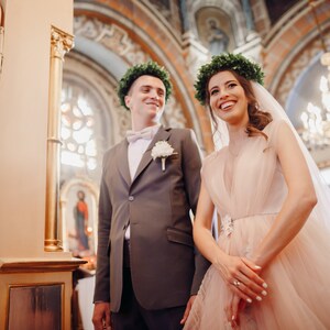 May include: A bride and groom in a church, the bride is wearing a blush pink dress with a veil and a green leaf crown, the groom is wearing a gray suit with a white button-up shirt and a green leaf crown.