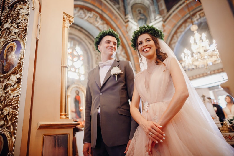 May include: A bride and groom in a church, the bride is wearing a blush pink dress with a veil and a green leaf crown, the groom is wearing a gray suit with a white button-up shirt and a green leaf crown.