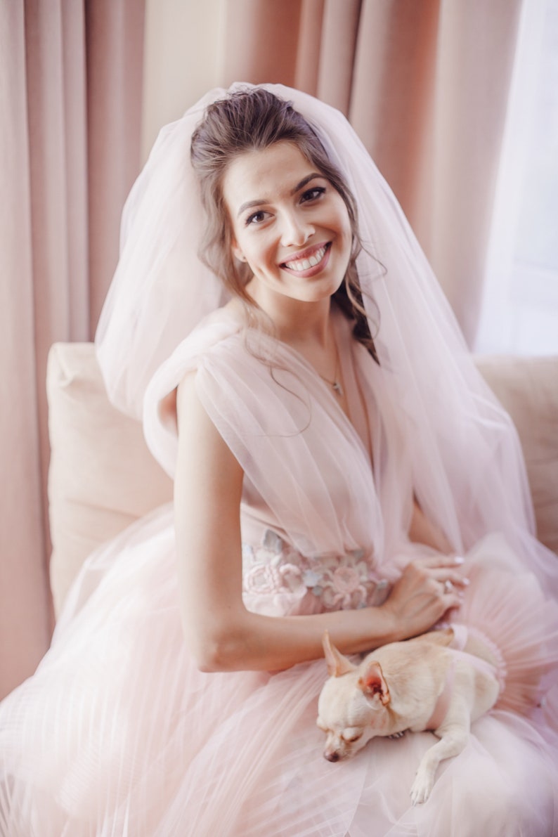 May include: A bride in a pink tulle wedding dress with a veil and a small chihuahua dog sitting on her lap.