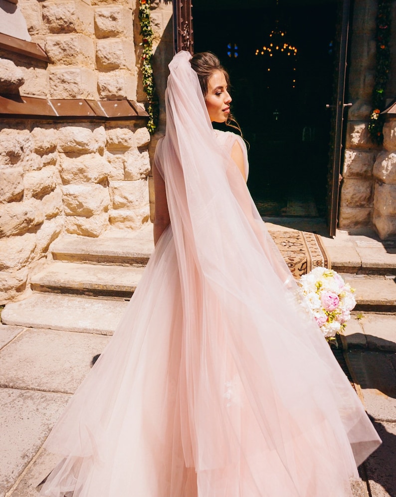 May include: A bride in a long, flowing pink tulle wedding dress with a veil. She is standing on the steps of a building with a bouquet of white and pink flowers in her hand.