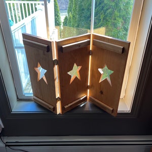 May include: A set of three wooden window shutters with cut-out star designs. The shutters are closed and are made of light brown wood.