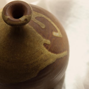 May include: A handcrafted ceramic vase with a rounded body and a narrow neck. The vase features a brown base with a lighter brown and tan design. The opening is a dark brown color. The vase is on a white surface.