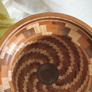 May include: A handcrafted wooden bowl with a spiral design. The bowl features a mosaic pattern of various wood tones, including light and dark browns, and beige. The interior has a dark brown center, and the rim has a decorative border.