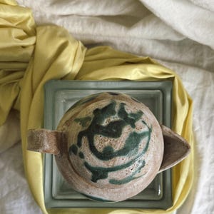 May include: A ceramic teapot with a lid, featuring a beige base and green abstract patterns. The teapot has a handle and a spout. It rests on a square glass block, with yellow fabric and white cloth in the background.