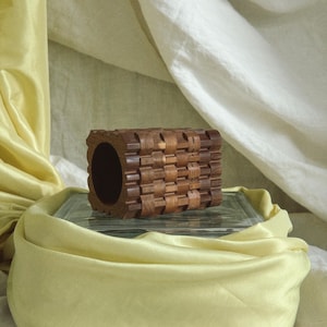May include: A handcrafted, cylindrical wooden object with a woven pattern. The dark brown wood has a smooth finish and a large central opening. The object is displayed on a glass block, with a yellow fabric draped around it.