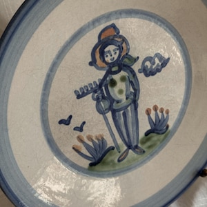 May include: Hand-painted ceramic plate with a blue and white design. The plate features a central illustration of a person in a hat, holding a rake, with birds and floral accents. The rim has a double blue stripe.