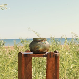 May include: A round, iridescent ceramic pot with horizontal ridges and a wide opening, resting on a tall, four-legged wooden stand. The pot features a mottled finish with shades of green, brown, and blue, while the stand is made of dark wood.