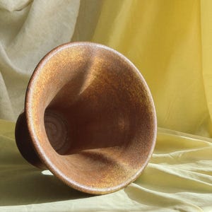 May include: A brown ceramic vase with a wide, flared opening and a rounded base. The vase has a textured surface and a warm, earthy color. The vase is set against a backdrop of yellow and cream-colored fabric.