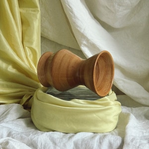 May include: A handcrafted wooden vase with a unique hourglass shape. The vase is made of light brown wood and sits atop a folded yellow cloth. The background features draped white and yellow fabric, creating a soft, textured backdrop.
