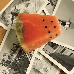 May include: A wedge-shaped soap bar designed to resemble a slice of watermelon, featuring a vibrant red interior, a green rind edge, and several small, dark, circular indentations scattered across the surface to represent seeds.