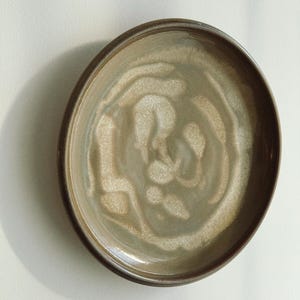 May include: A ceramic plate with a circular shape and a dark brown rim. The interior of the plate features a swirling pattern in shades of beige and light brown, creating a textured appearance. The plate is designed for serving food.
