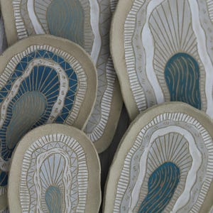 Pode incluir: Uma coleção de pratos de cerâmica com um design de concha de ostra. Os pratos apresentam uma base bege com detalhes em branco, cinza e azul-petróleo. Têm uma forma orgânica única e estão dispostos de forma atraente.