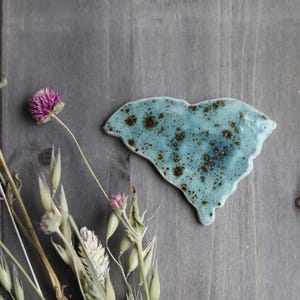 May include: A ceramic piece in the shape of the state of South Carolina, with a speckled turquoise glaze. Dried flowers and foliage, including purple clover, are arranged on a weathered wooden surface.