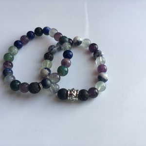 May include: Two beaded bracelets with a silver charm. The bracelets are made with amethyst, green aventurine, blue lapis lazuli, fluorite, and lava rock beads.