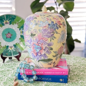 May include: A yellow ceramic ginger jar with a lid, adorned with a floral design in shades of pink, purple, and blue. The jar is set on a stack of books, with a beaded necklace in the foreground. A decorative plate is visible in the background.