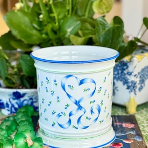 Op de afbeelding: Een witte keramische plantenbak met een blauwe rand en basisafwerking. De plantenbak heeft een blauw lint hart ontwerp en kleine blauwe bloemaccenten. De plantenbak staat op een boek met de woorden "Sweet Floral Life".