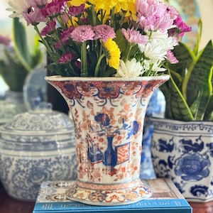 May include: A decorative porcelain vase with a floral design in blue, orange, and white, filled with a vibrant bouquet of yellow, pink, and white chrysanthemums. The vase sits atop a stack of books.
