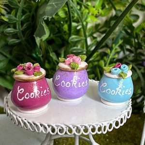 Könnte beinhalten: Drei Miniatur-Keksdosen aus Keramik mit floralen Akzenten. Jede Dose ist in einer anderen Farbe bemalt: rosa, lila und blau. Die Dosen sind mit "Cookies" in weißer Schrift beschriftet.