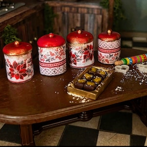 Könnte beinhalten: Vier rote und weiße Keramikdosen mit einem Weihnachtsstern-Design, die auf einem Holztisch mit einem goldenen Tablett mit Schokoladenbonbons stehen.