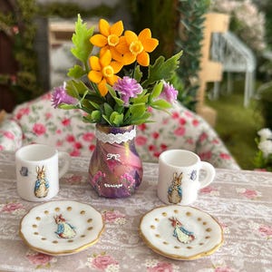 Könnte beinhalten: Miniatur-Teeservice mit einer Vase mit orangefarbenen und lila Blumen, zwei weißen Tassen und zwei kleinen Tellern. Die Tassen und Teller sind mit einer Kaninchenillustration und goldenen Verzierungen verziert. Die Vase hat ein Vintage-Design.