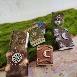 Könnte beinhalten: Sechs Miniaturbücher mit verschiedenen Einbänden, darunter Braun, Grün und Silber. Jedes Buch hat ein Halbmond-Design und einige haben dekorative Verzierungen wie einen türkisfarbenen Stein und ein silbernes Herz.