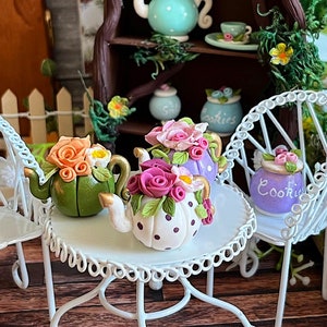 Könnte beinhalten: Drei Miniatur-Teekannen, verziert mit Blumen und Punkten, stehen auf einem weißen Tisch mit einem weißen Stuhl. Die Teekannen sind aus Keramik und haben einen verspielten, märchenhaften Garten-Look.