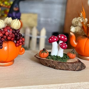 Könnte beinhalten: Eine Miniatur-Szene mit Pilzen und Kürbissen mit drei roten und weißen Pilzen auf einer Holzscheibe mit grünem Moos. Zwei kleine orangefarbene Kürbisse befinden sich auf beiden Seiten der Pilze.