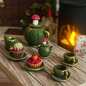 Könnte beinhalten: Miniatur-Teeservice in Grün mit goldenen Akzenten, einschließlich einer Teekanne, einem Sahnekännchen, zwei Tassen und zwei Cupcakes. Die Teekanne und das Sahnekännchen sind mit pilzförmigen Stöpseln versehen. Die Cupcakes sind mit Früchten und Zuckerguss verziert.