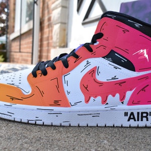 May include: White high-top sneakers with a black swoosh and a pink, orange, and yellow paint design. The sneakers have black laces and the word 'AIR' is written on the side.