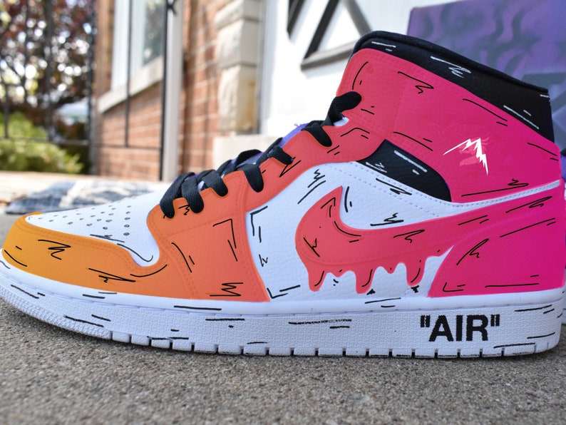 May include: White high-top sneakers with a black swoosh and a pink, orange, and yellow paint design. The sneakers have black laces and the word 'AIR' is written on the side.