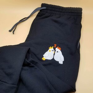 Può includere: Pantaloni della tuta neri con vita con coulisse. I pantaloni presentano un ricamo di due uccelli bianchi con dettagli gialli e arancioni e piccoli cuori rossi sopra. I pantaloni sono realizzati in materiale morbido.