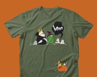 May include: Olive green t-shirt featuring a Halloween-themed graphic. The design includes cartoon birds in costumes, ghosts, and a pumpkin with a fox. A speech bubble says "booo".