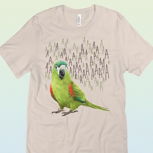 Puede incluir: Una camiseta beige con una ilustración de un loro verde y rojo, y la palabra "AAAA" repetida muchas veces en un patrón.