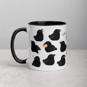 Könnte beinhalten: Weiße Keramiktasse mit schwarzem Rand und Henkel. Die Tasse zeigt ein Muster aus schwarzen Comic-Rabenvögeln mit verschiedenen Gesichtsausdrücken. Der Text "CAW" ist auf der rechten Seite der Tasse sichtbar.